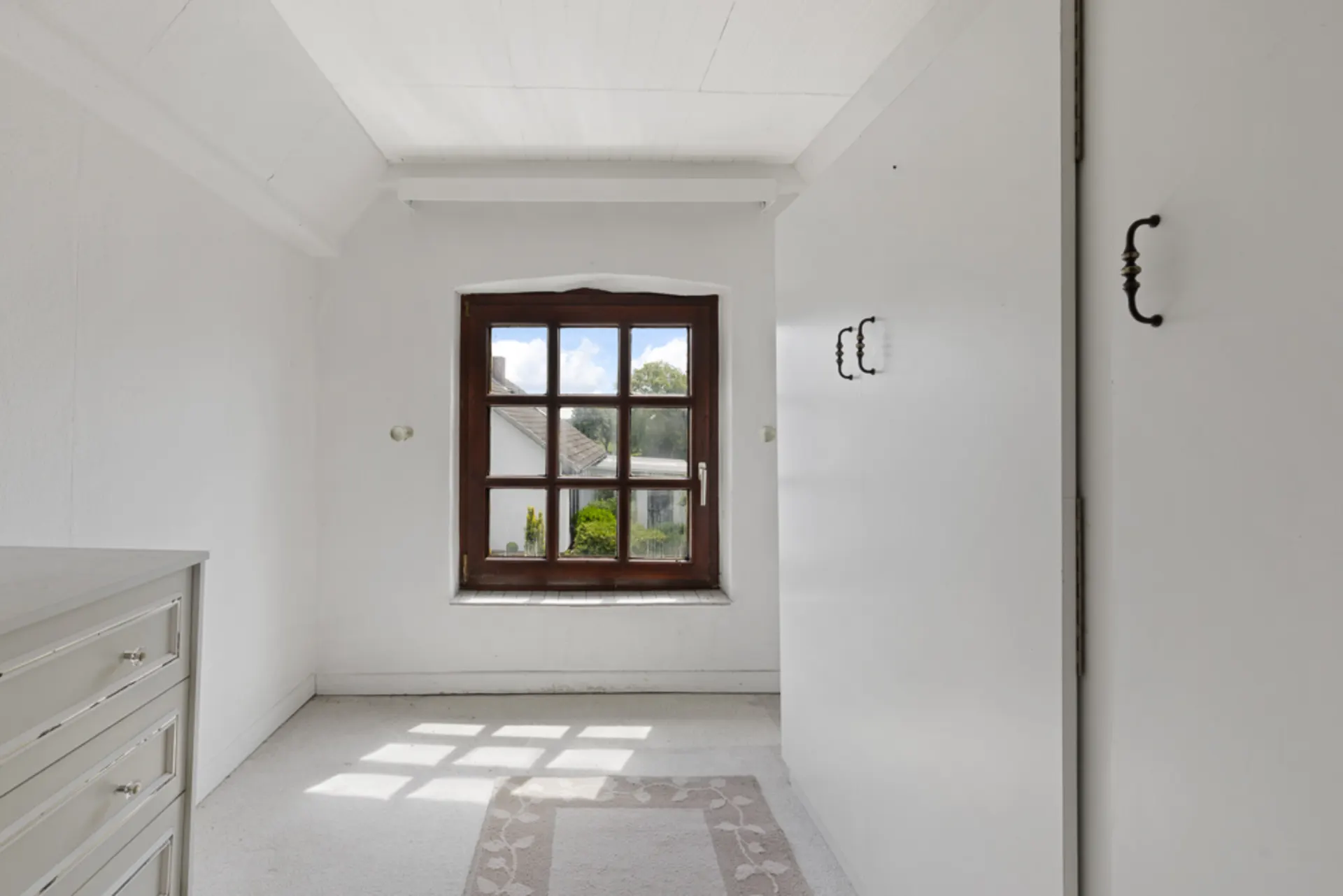 Helles Zimmer mit weißem Teppich, Fenster mit Holzrahmen, Schrankwand und Kommode.