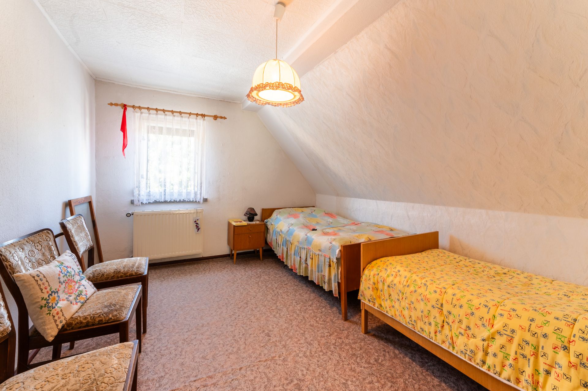 Schlafzimmer mit zwei Einzelbetten, drei Stühlen und kleinem Fenster mit Gardine.