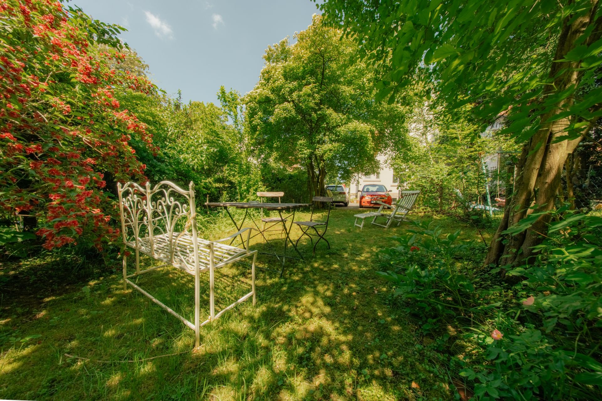 Grüner Garten mit Metallbank, Tisch, Stühlen und Bäumen im Hintergrund.