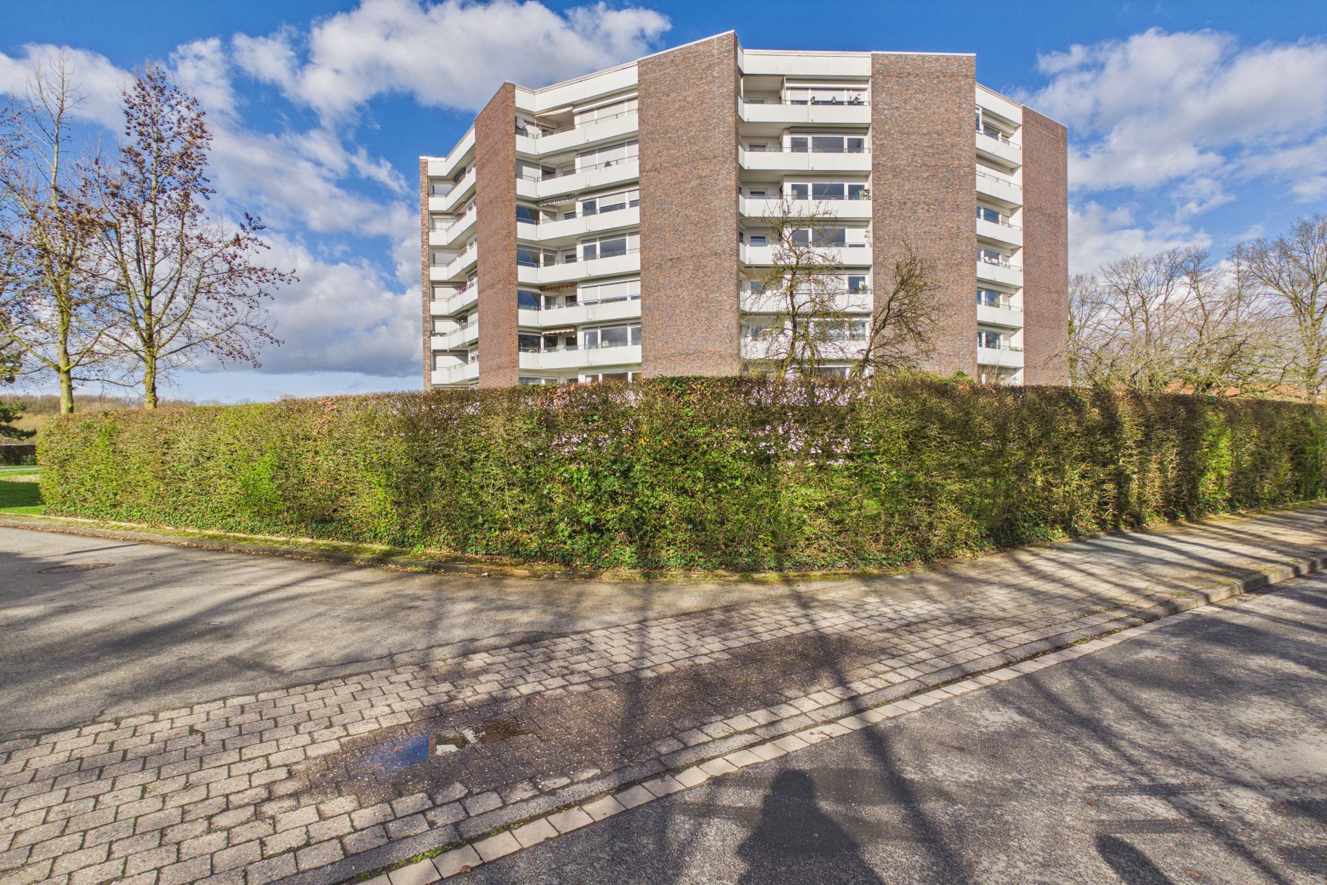 Mehrstöckiges Wohnhaus mit Balkonen und Hecke im Vordergrund bei sonnigem Wetter.