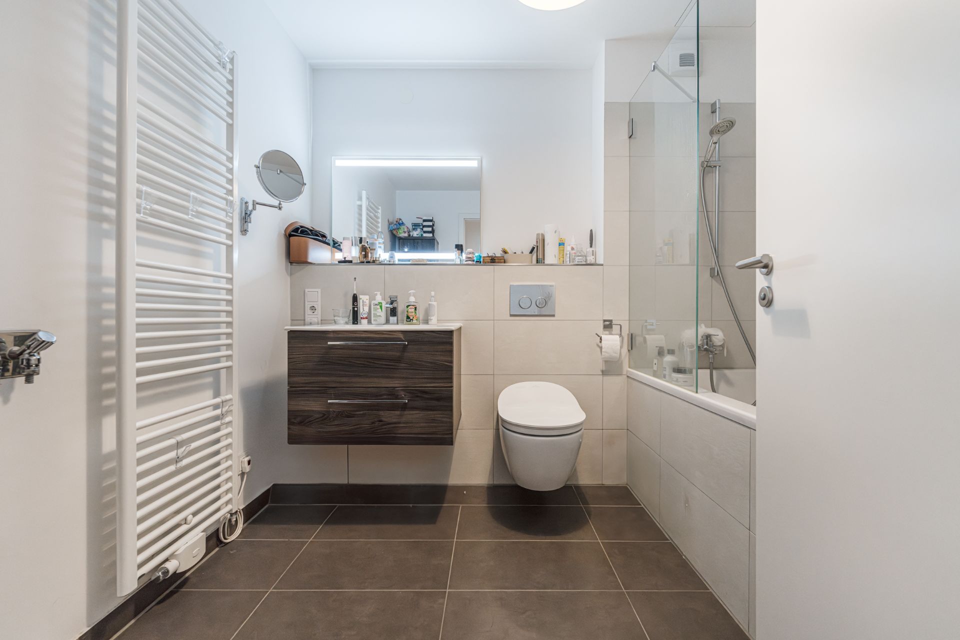 Modernes Badezimmer mit Wandtoilette, Waschbecken, Spiegel und Badewanne mit Glasabtrennung.