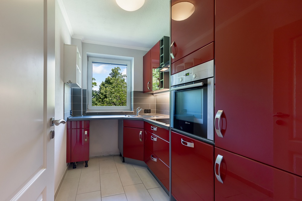 Moderne Küche mit roten Hochglanzschränken, Einbaugeräten und Fenster mit Blick ins Grüne.