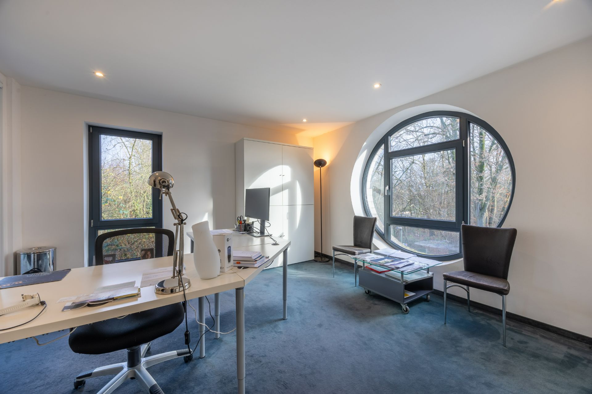 Helles Arbeitszimmer mit großem rundem Fenster, Schreibtisch, Stühlen und Schrank.