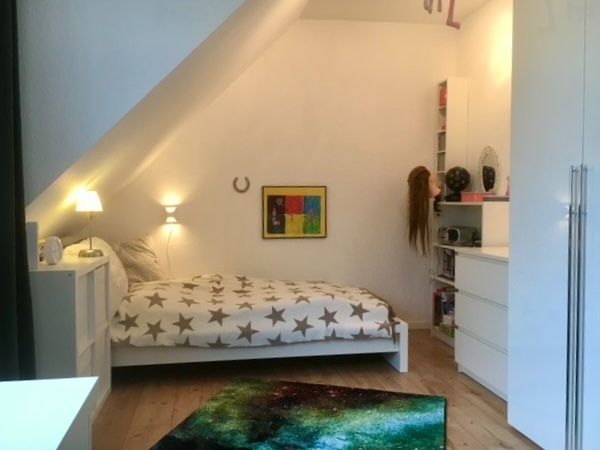 Schlafzimmer mit Bett, bunter Decke, Teppich, Kommode und Wandbild unter Dachschräge.