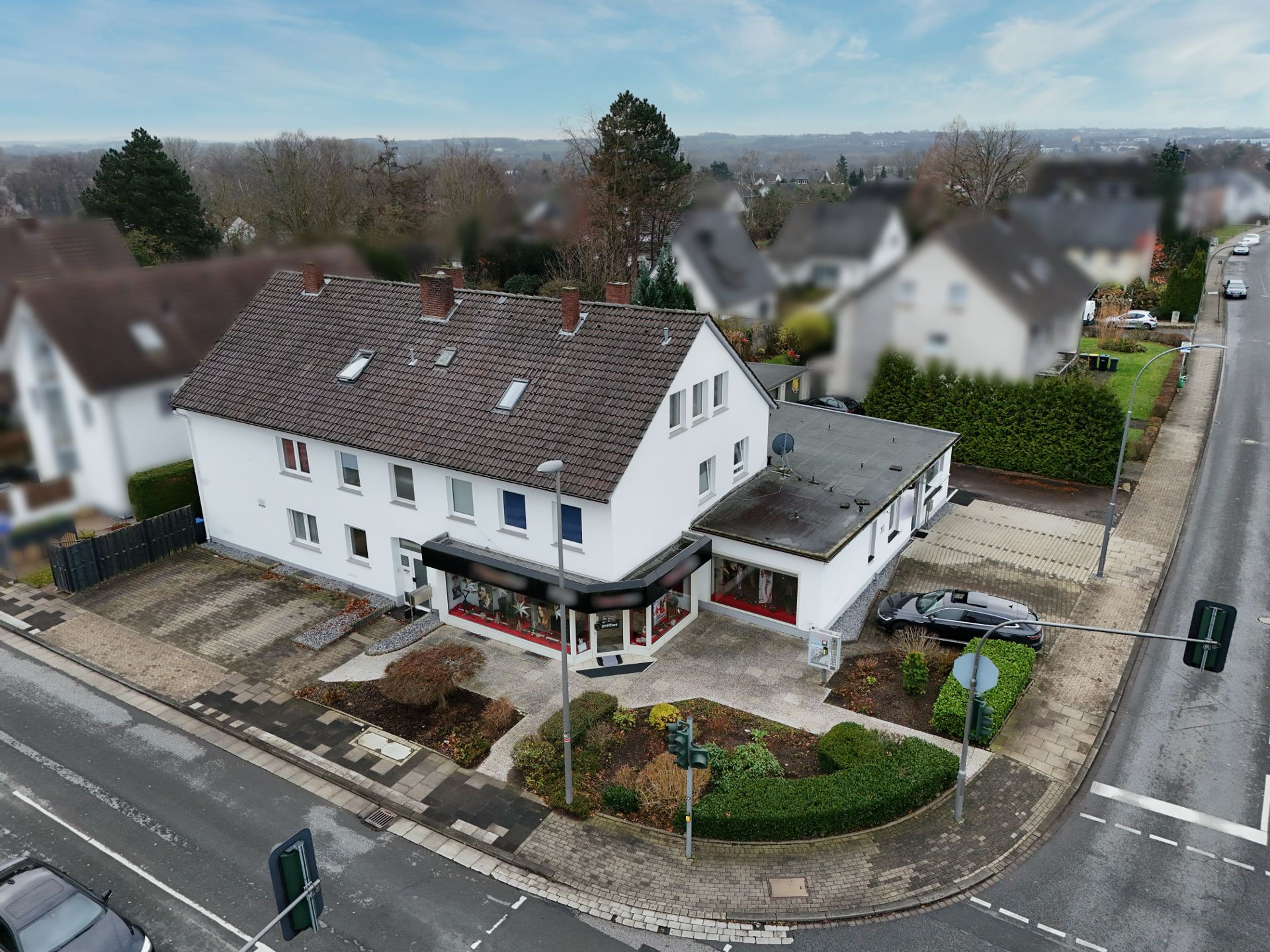 Eckhaus mit Schaufenstern, Parkplatz und gepflegtem Vorgarten an einer Kreuzung.