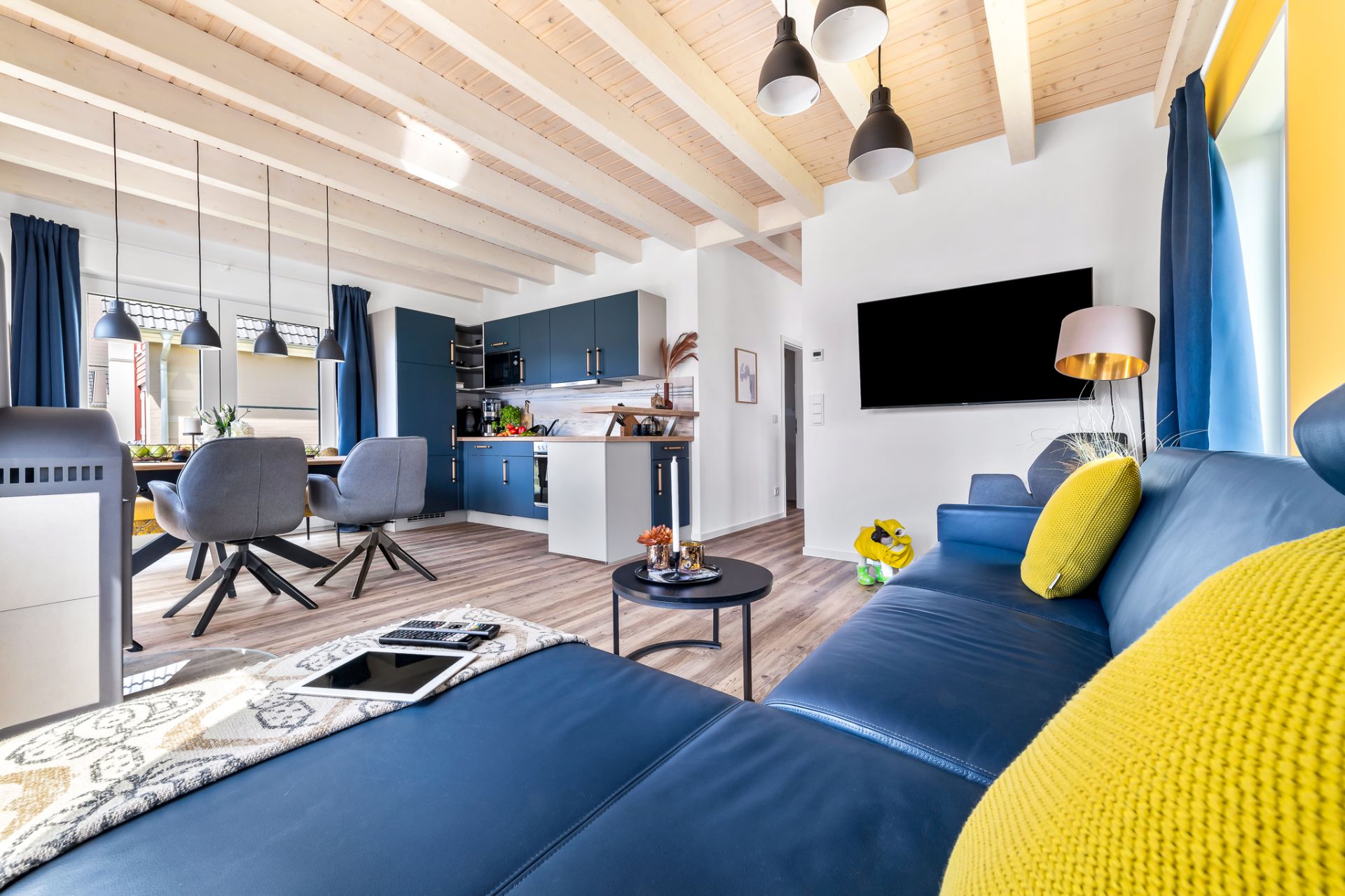 Modernes Wohnzimmer mit blauer Eckcouch, Fernseher, Esstisch und offener Küche.