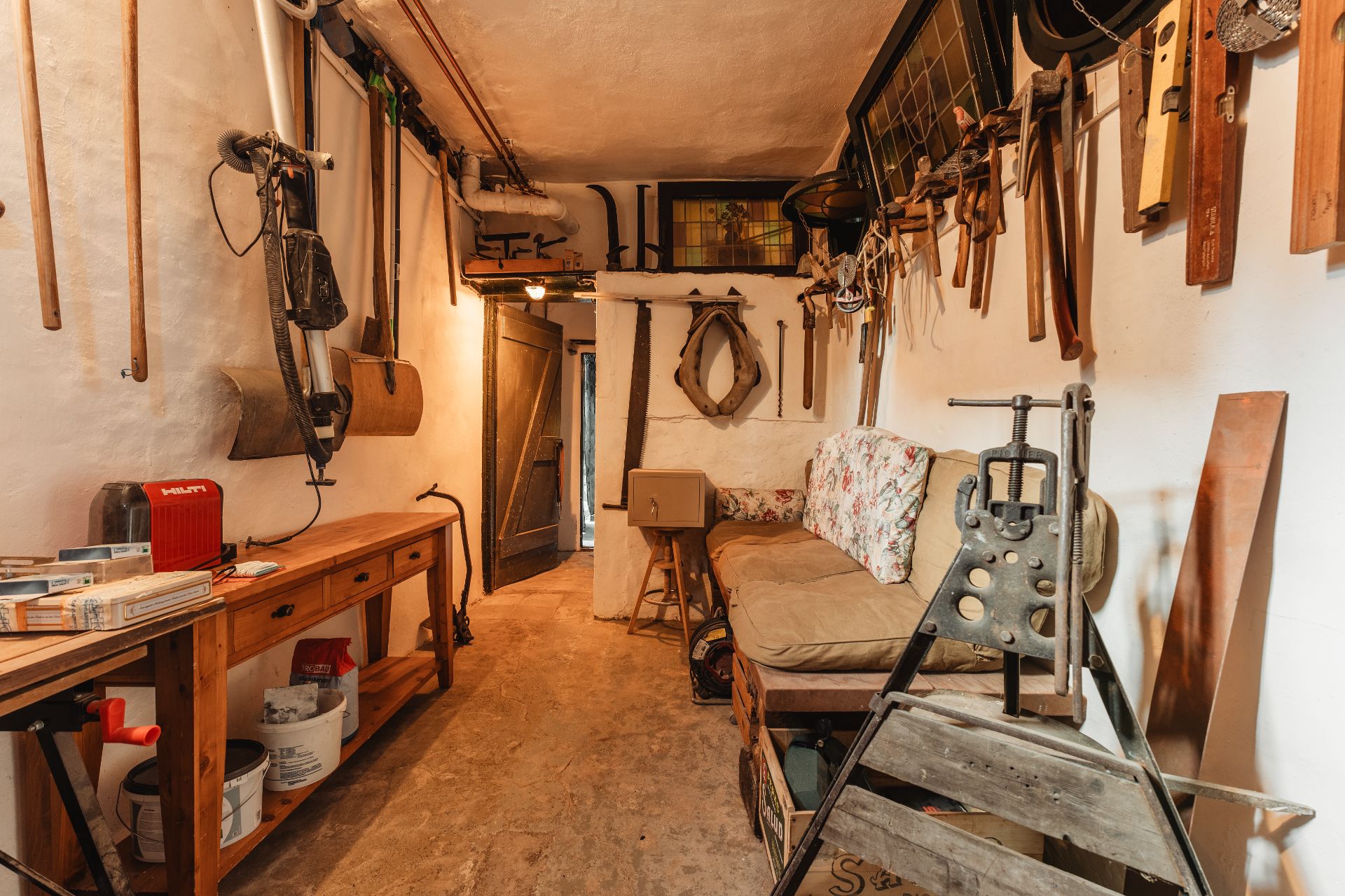 Werkstattraum mit vielen Werkzeugen an den Wänden, Holztisch, Sofa und Maschinen.