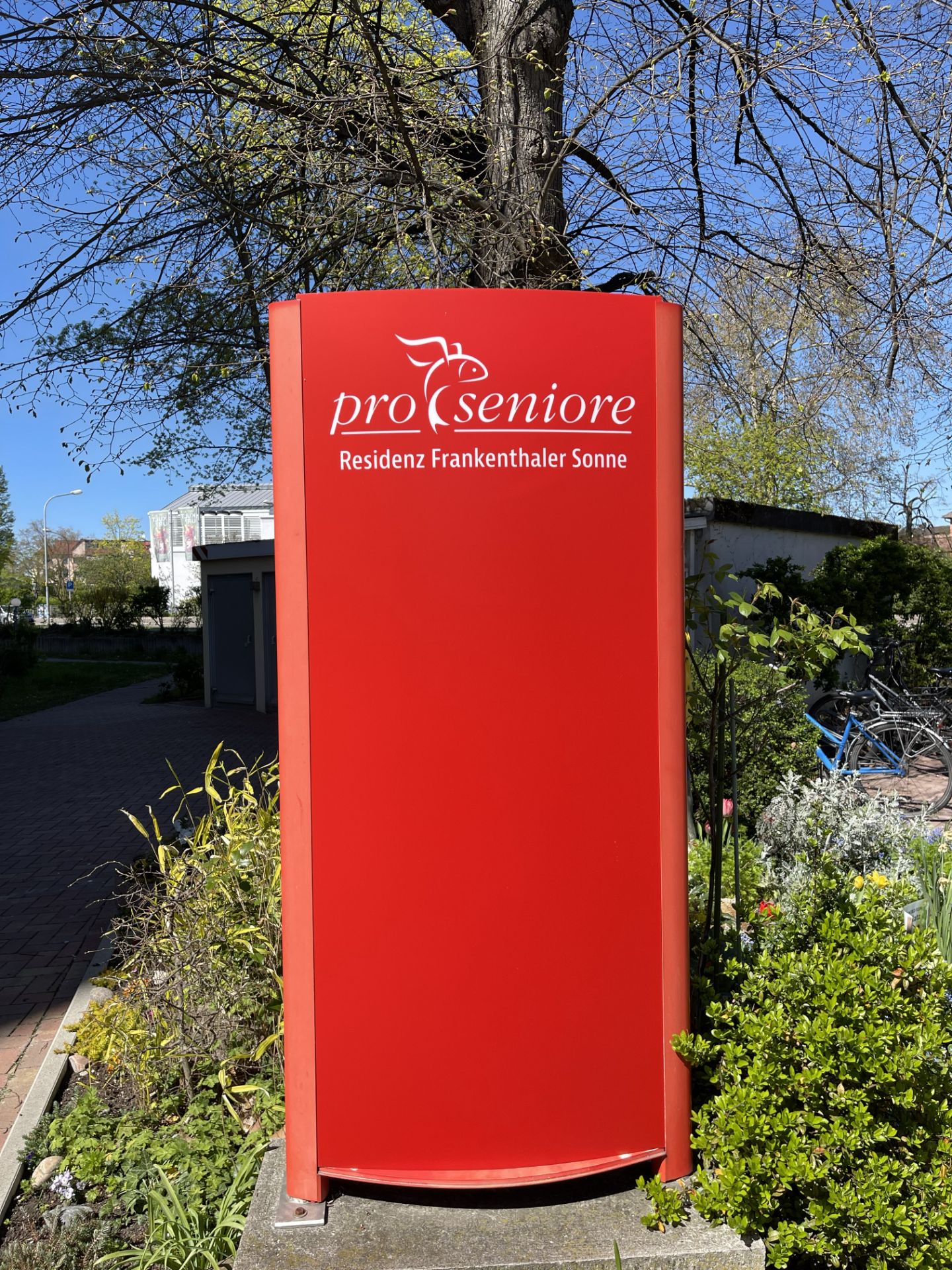 Rotes Schild mit weißer Aufschrift 'pro seniore Residenz Frankenthaler Sonne' in einem Garten.