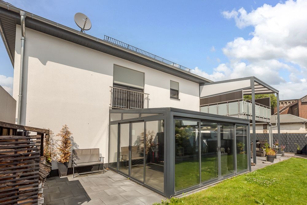 Moderne Rückseite eines Wohnhauses mit Wintergarten und Balkon, angrenzender Garten mit Terrasse.