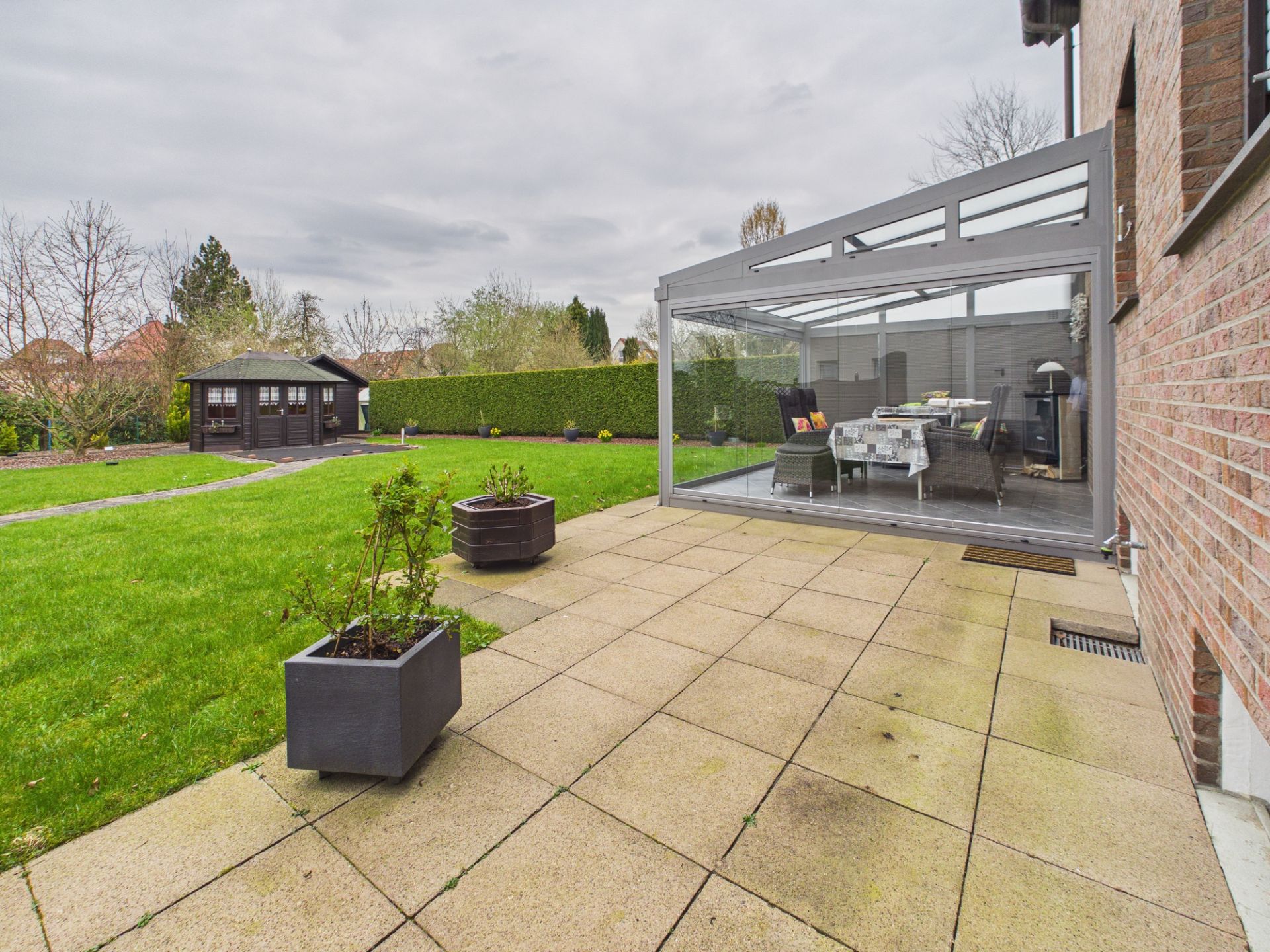 Große Terrasse mit Pflanzkübeln, Wintergarten und Blick auf Gartenhaus im grünen Garten.