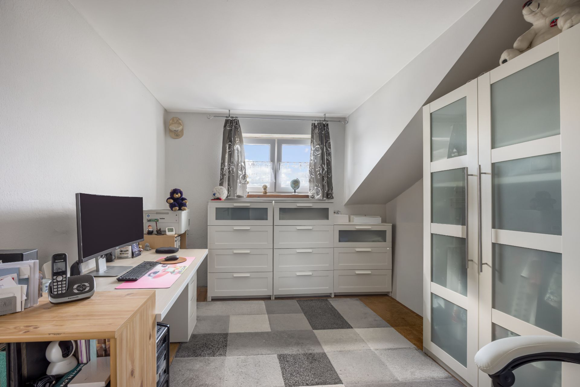 Helles Arbeitszimmer mit Schreibtisch, Computer, Regalen, Kommoden und Fenster mit Vorhängen.