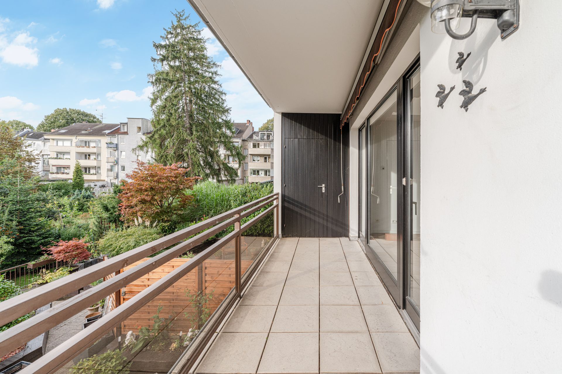 Balkon mit Fliesenboden, Glasgeländer und Blick auf Garten und Nachbargebäude.