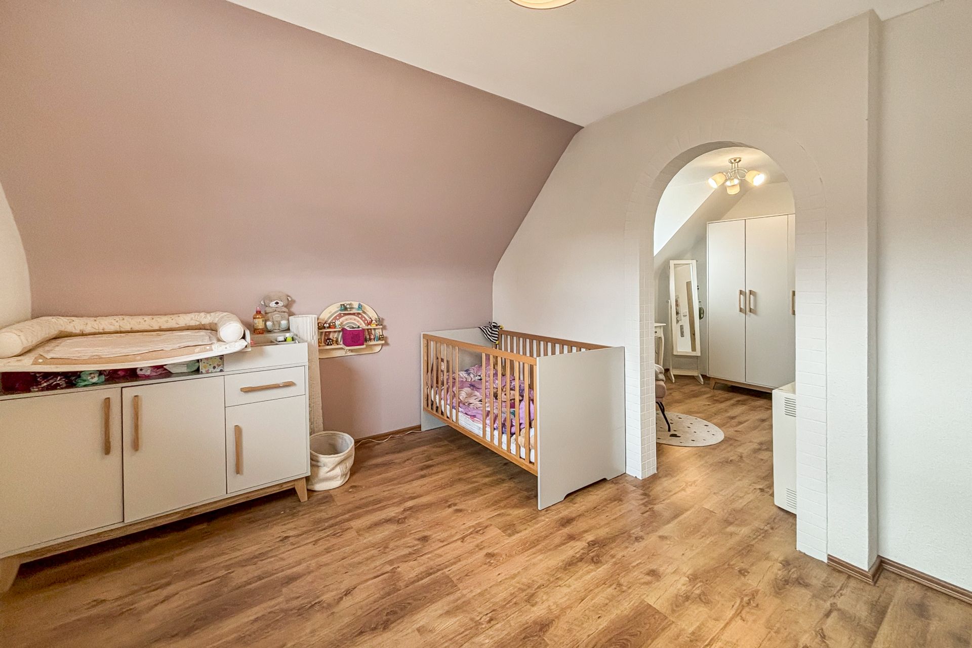Helles Kinderzimmer mit Holzboden, Babybett, Wickelkommode und Durchgang zu weiterem Raum.