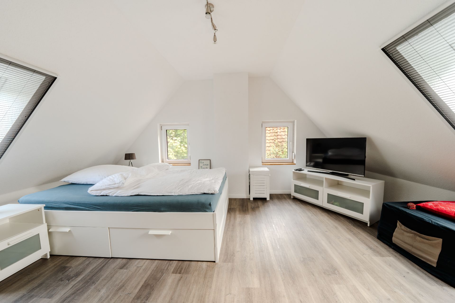 Helles Schlafzimmer mit Doppelbett, TV, Nachttischen und schrägen Fenstern im Dachgeschoss.