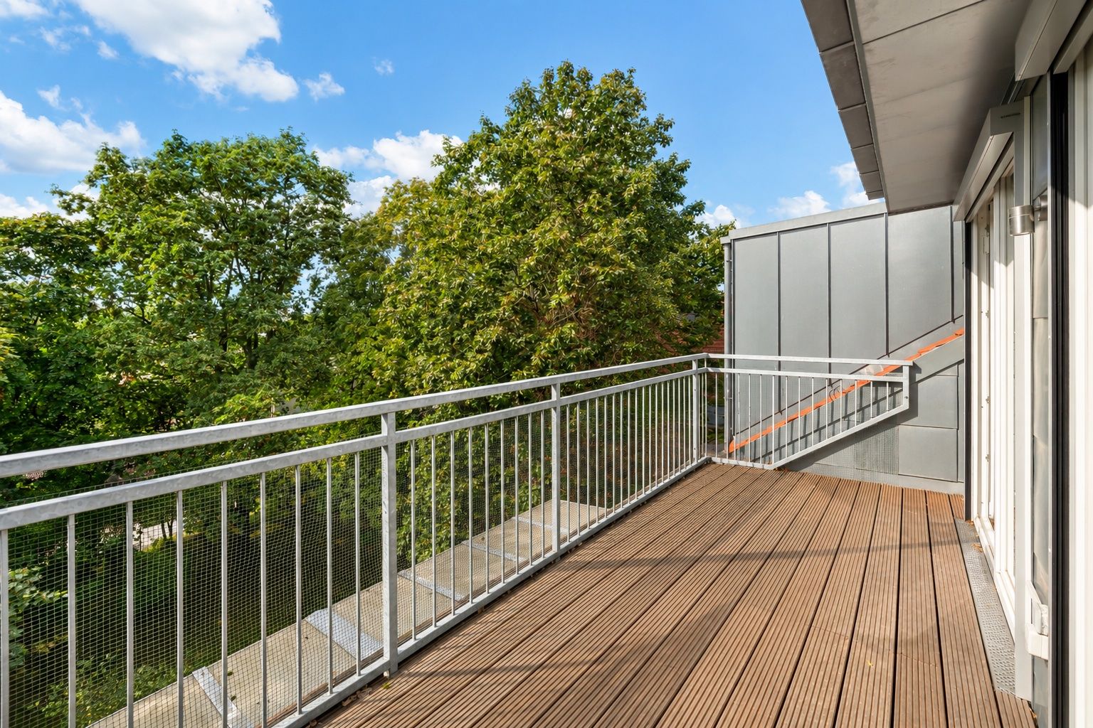 Großer Balkon mit Holzboden, Metallgeländer und Blick auf grüne Bäume.