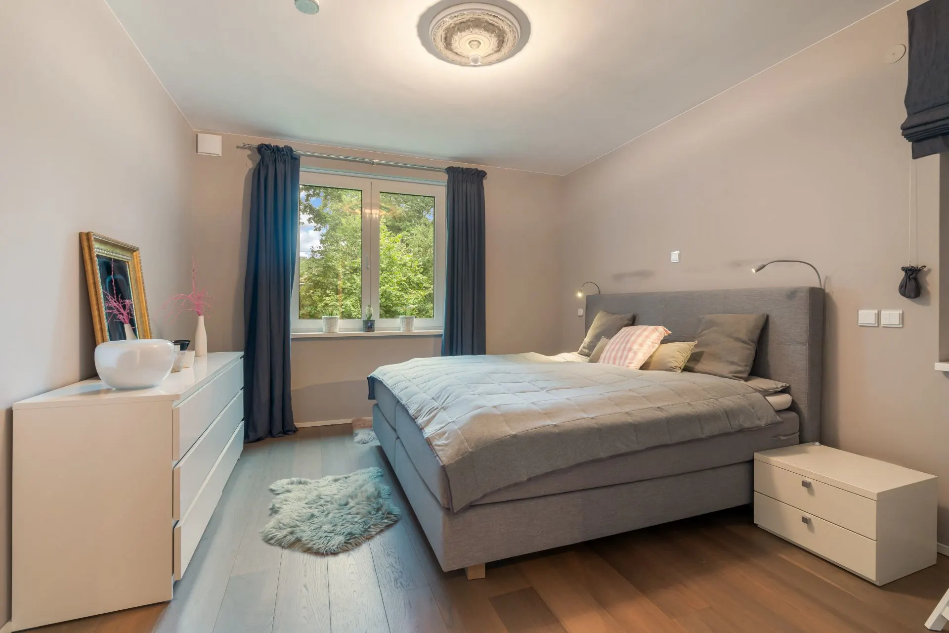Modernes Schlafzimmer mit Doppelbett, Kommode, Nachttisch und großem Fenster mit Vorhängen.