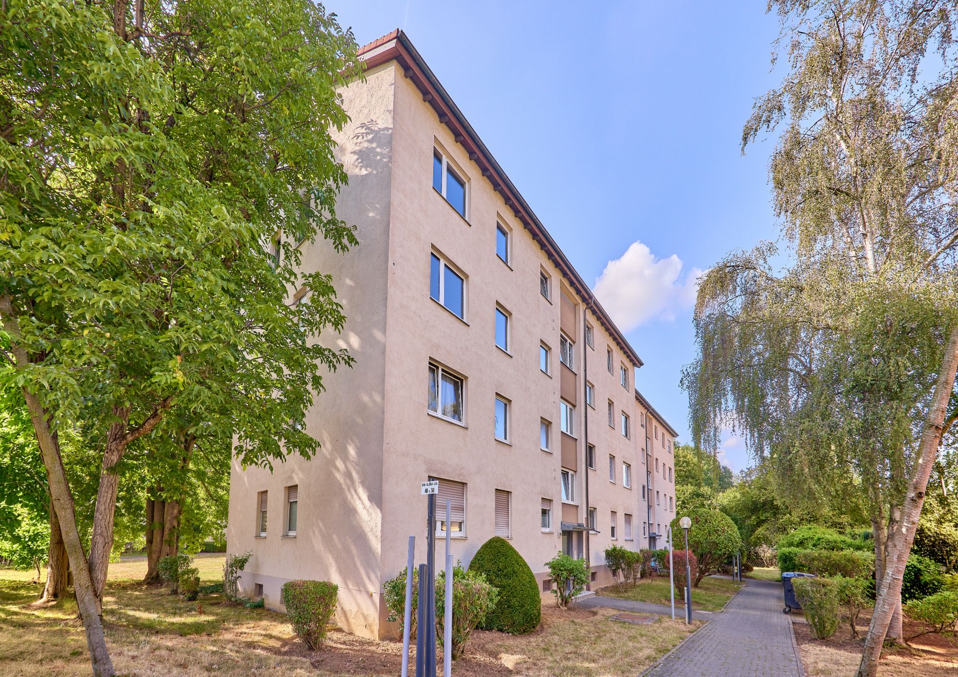 Vierstöckiges Wohnhaus mit mehreren Fenstern, umgeben von Bäumen und Gehweg.