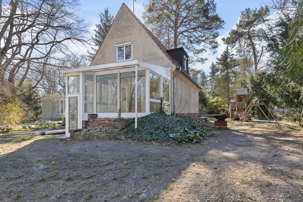Haus mit Wintergarten und Garten, Spielplatz im Hintergrund, umgeben von Bäumen.