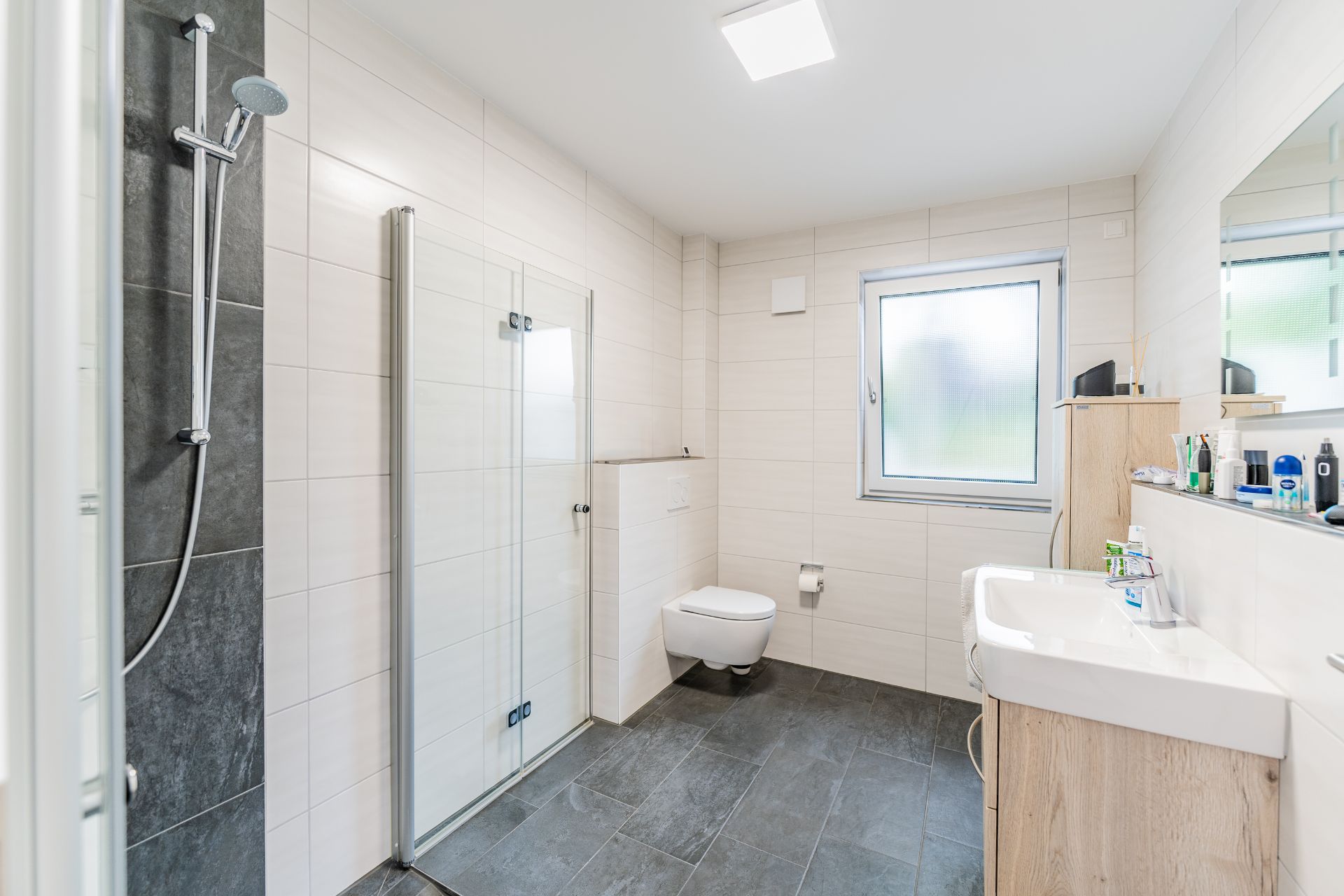 Modernes Badezimmer mit bodengleicher Dusche, WC, Fenster und Waschbecken mit Spiegel.