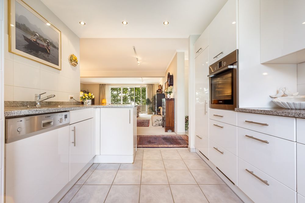 Moderne weiße Küche mit Einbaugeräten und Blick ins helle Wohnzimmer mit großen Fenstern.