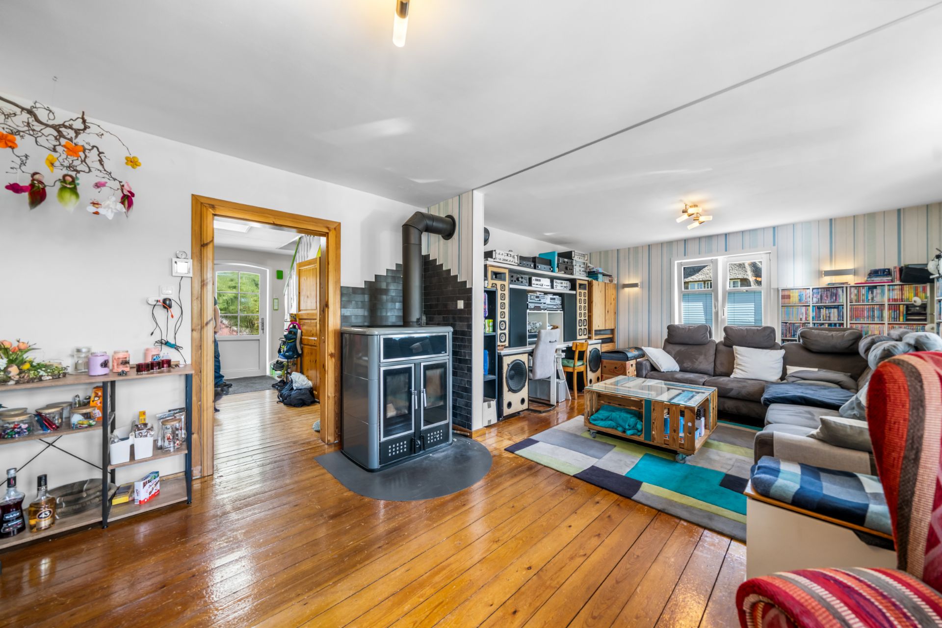 Wohnzimmer mit Holzfußboden, Kaminofen, Sofa, Regalen und offener Verbindung zur Küche.