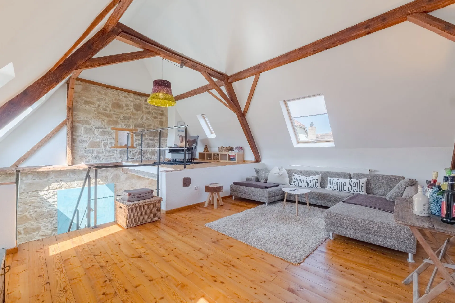Helles Wohnzimmer mit Holzboden, Ecksofa, Dachbalken und Steinwand unter Dachschrägen.