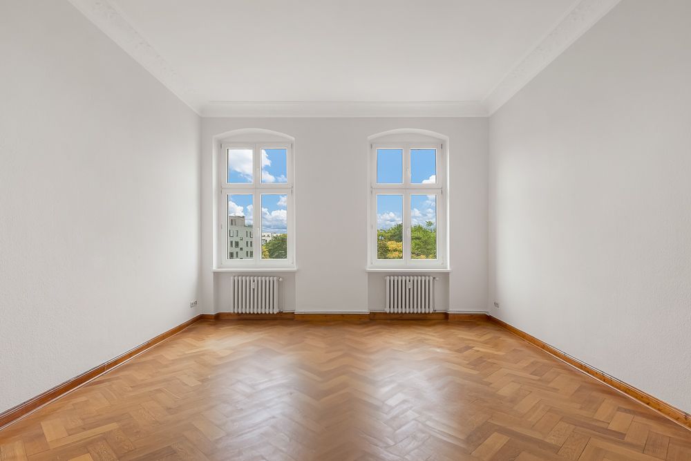 Leerer Raum mit Parkettboden, weißen Wänden und zwei großen Fenstern mit Heizkörpern.