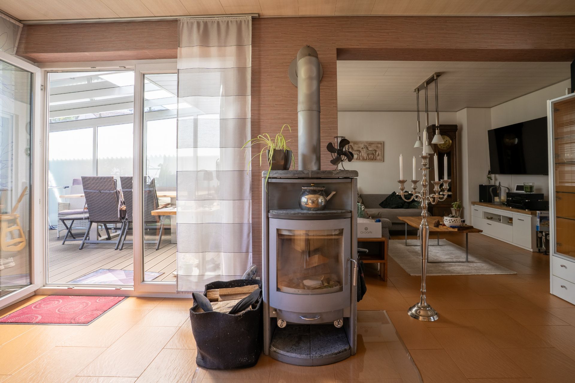 Wohnzimmer mit Kaminofen, Holzboden und Blick auf eine Terrasse mit Sitzgruppe.