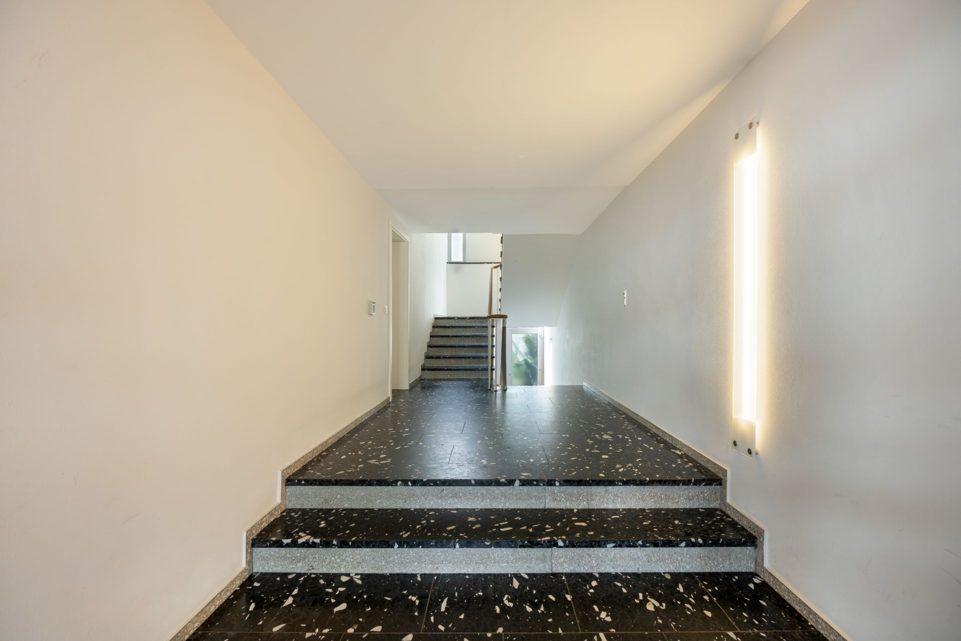 Heller Flur mit dunklem Bodenbelag und Treppe, weiße Wände mit langer Wandlampe.