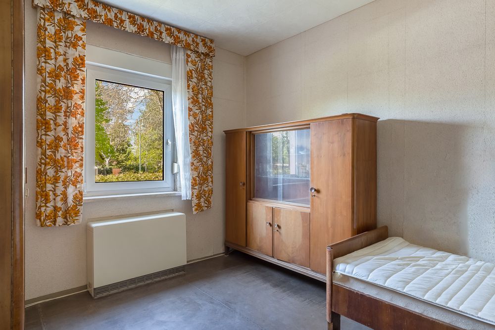 Kleines Zimmer mit Holzbett, Schrank und Fenster mit gemusterten Vorhängen.