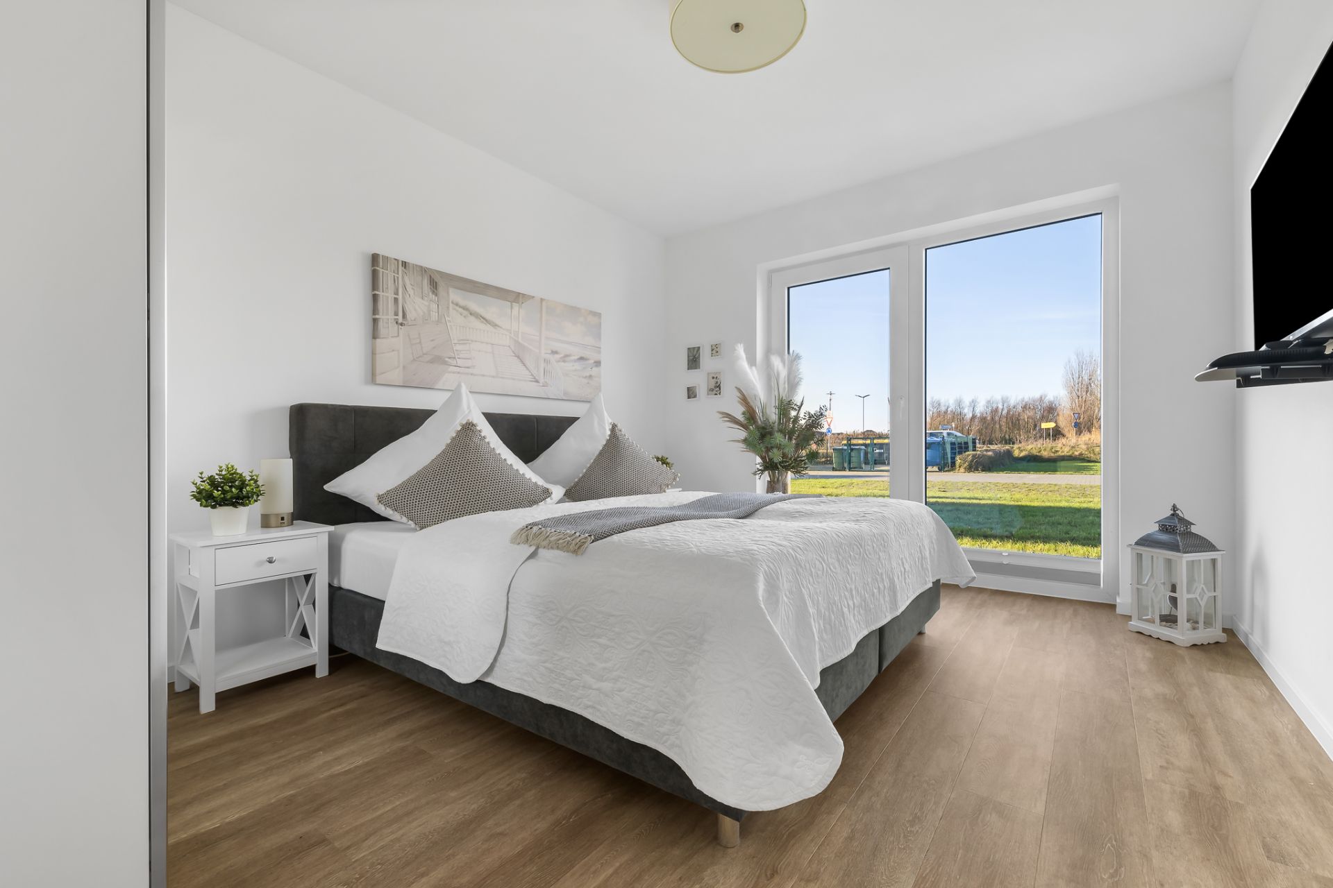 Helles Schlafzimmer mit Doppelbett, großem Fenster, Nachttisch und Fernseher an der Wand.