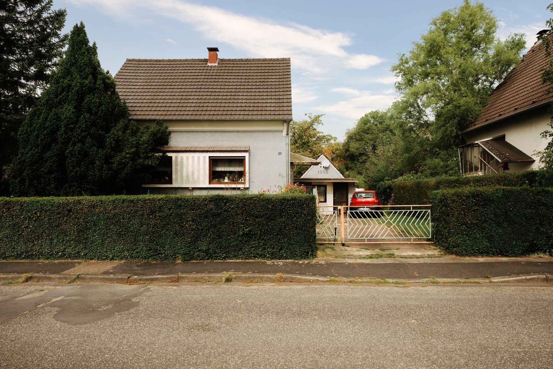 Vorderansicht eines Hauses mit Gartenzaun, Hecke und geparktem Auto in der Einfahrt.