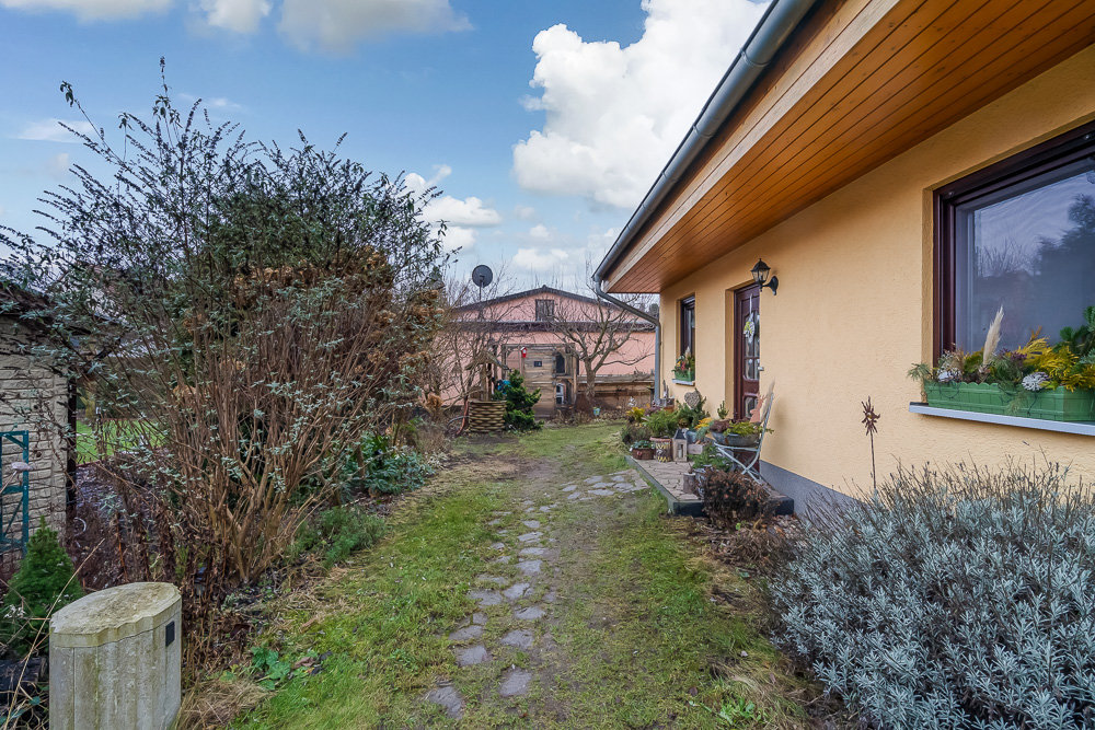 Gartenweg mit Pflanzen neben einem gelben Wohnhaus, im Hintergrund weiteres Gebäude.