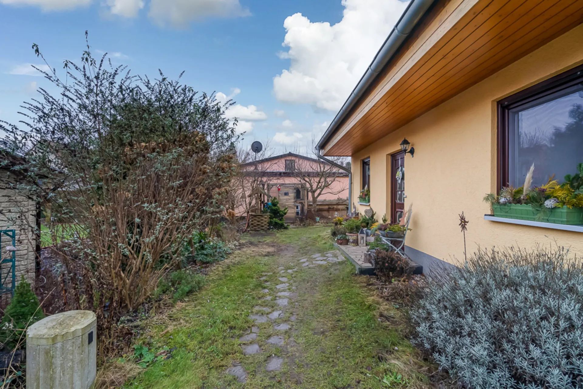 Gartenweg mit Pflanzen neben einem gelben Wohnhaus, im Hintergrund weiteres Gebäude.