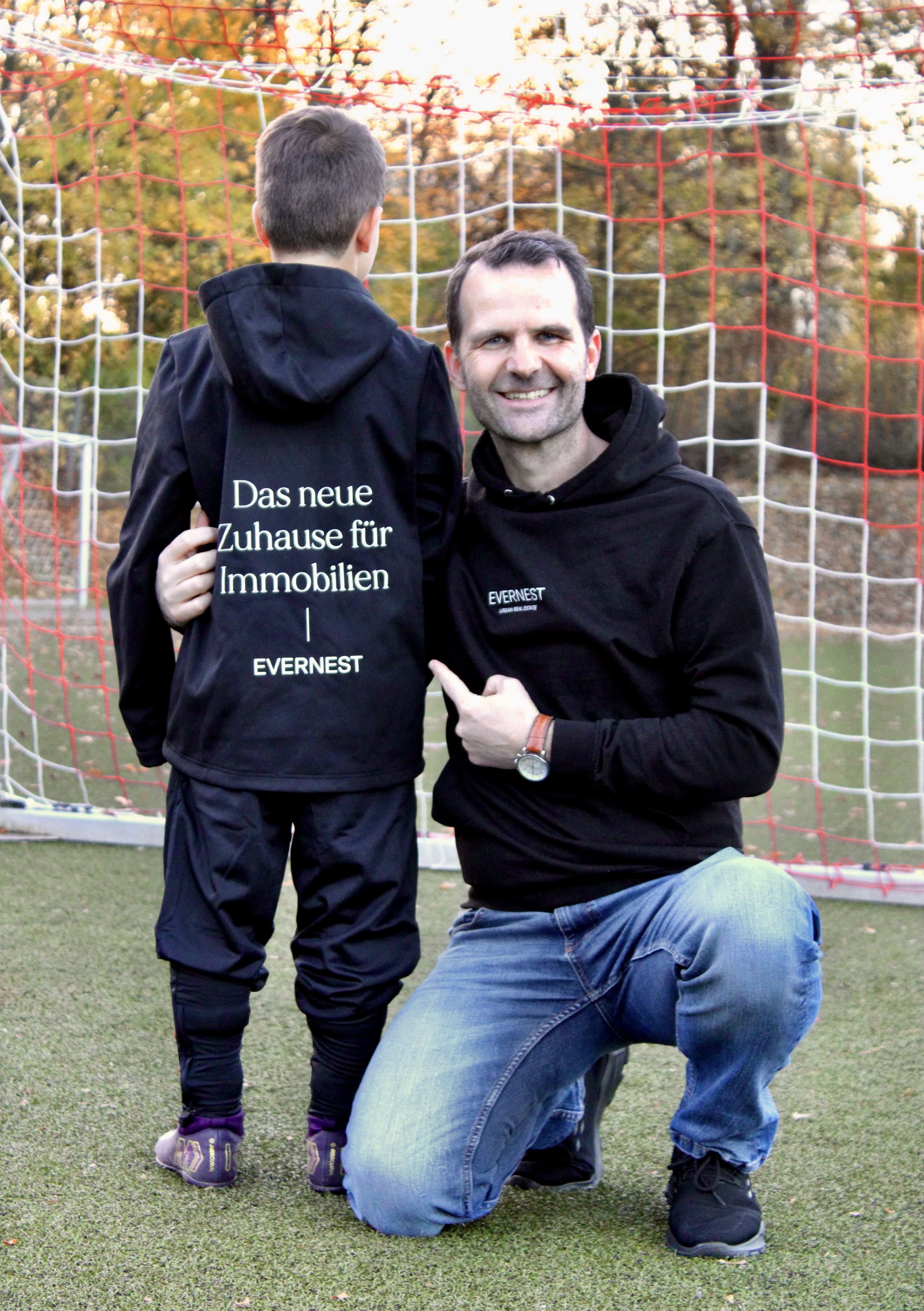 Ein Mann und ein Kind stehen vor einem Fußballtor. Beide tragen schwarze Hoodies mit Evernest-Logo.