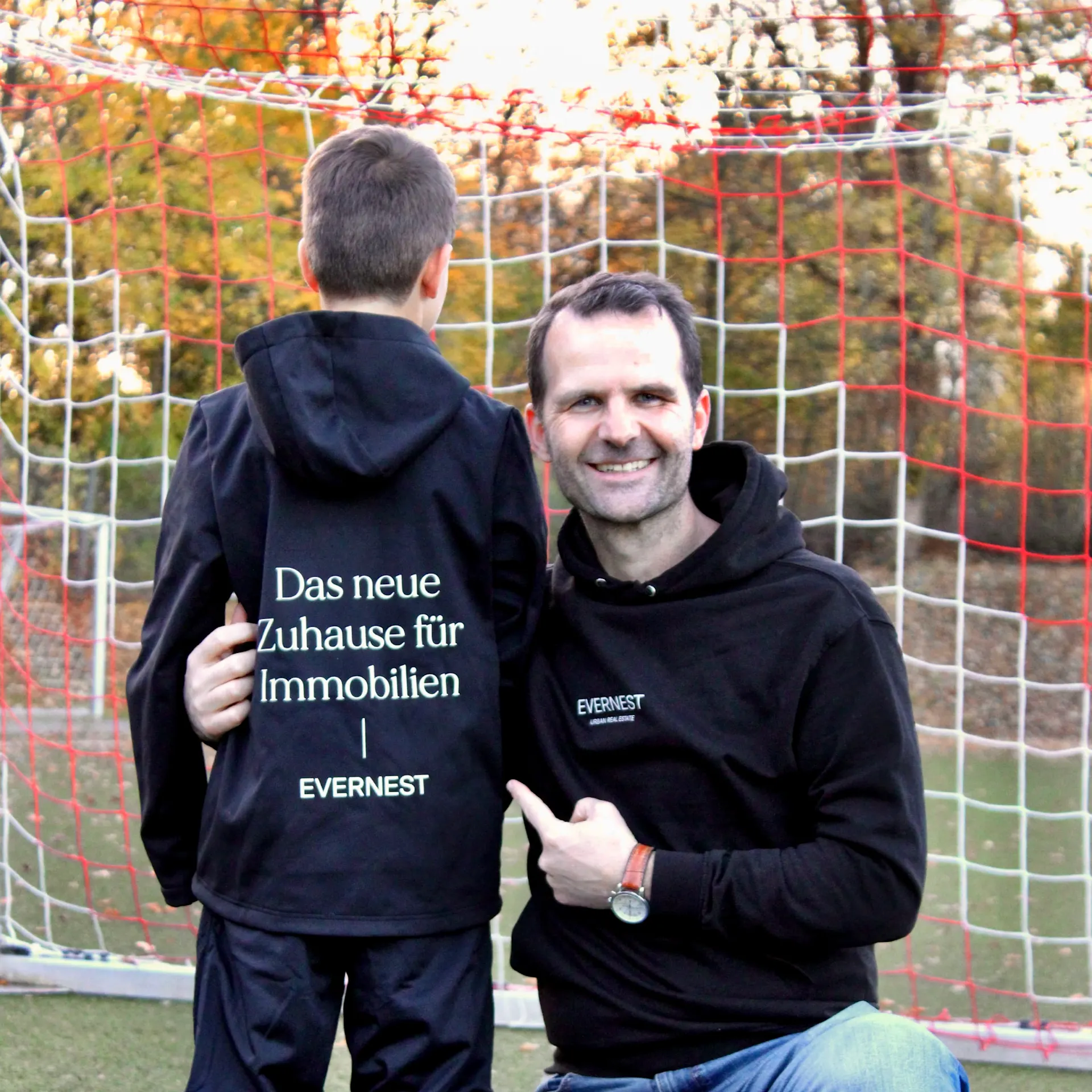 Ein Mann und ein Kind stehen vor einem Fußballtor. Beide tragen schwarze Hoodies mit Evernest-Logo.