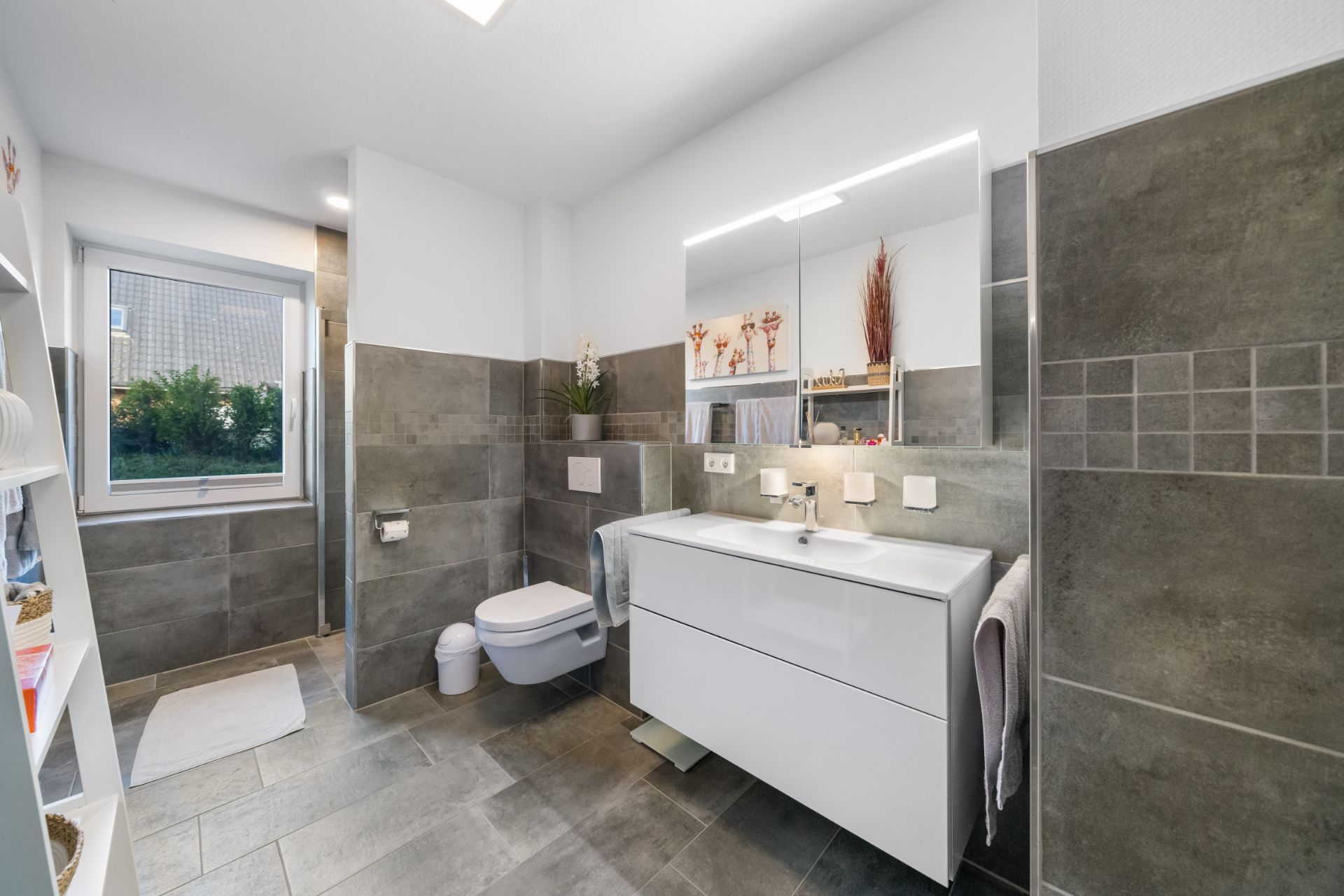 Modernes Badezimmer mit großem Spiegel, weißem Waschbecken und grauen Fliesen.
