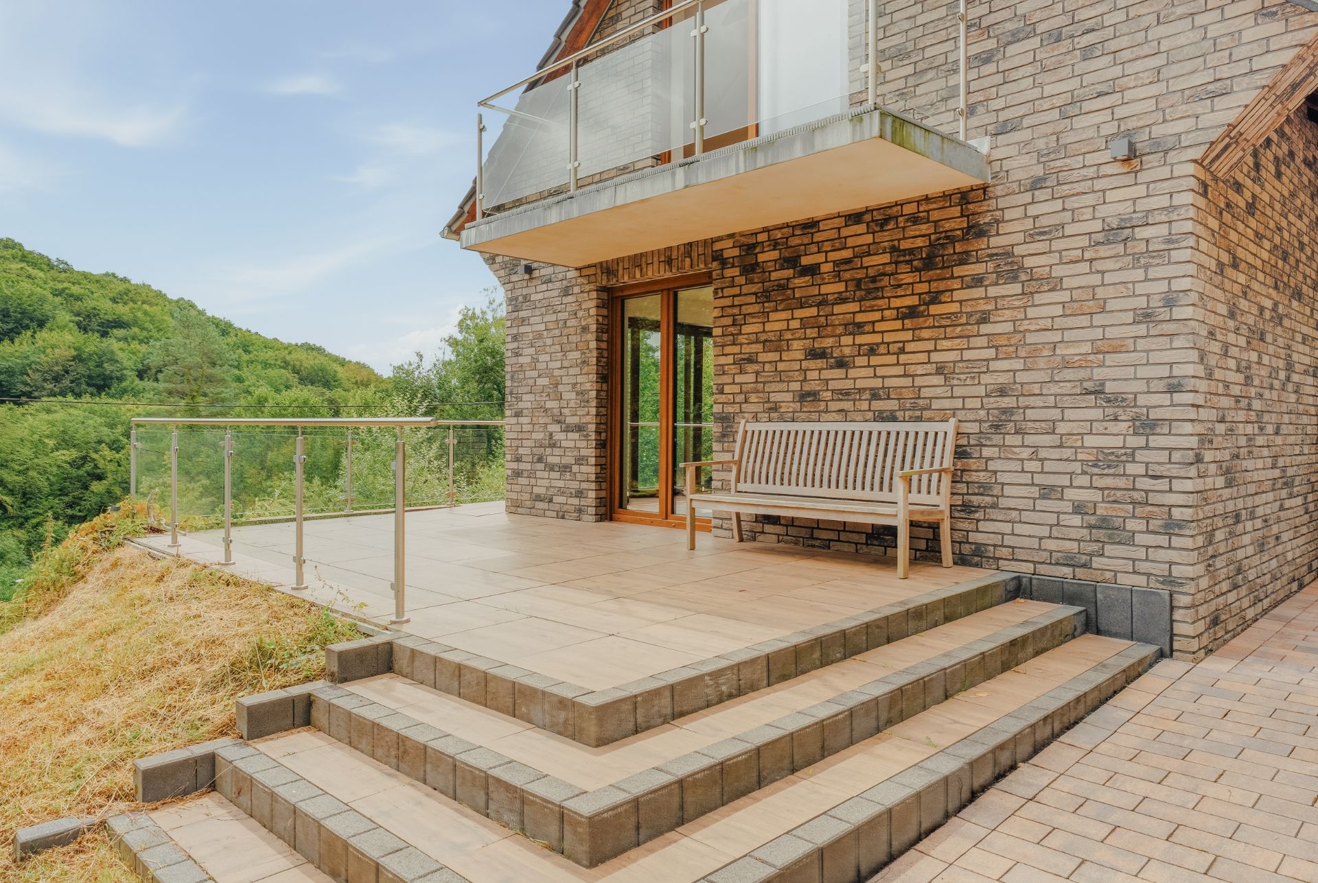 Geflieste Terrasse mit Holzbank, Glaskgeländer und Treppe an einem Backsteinhaus im Grünen.