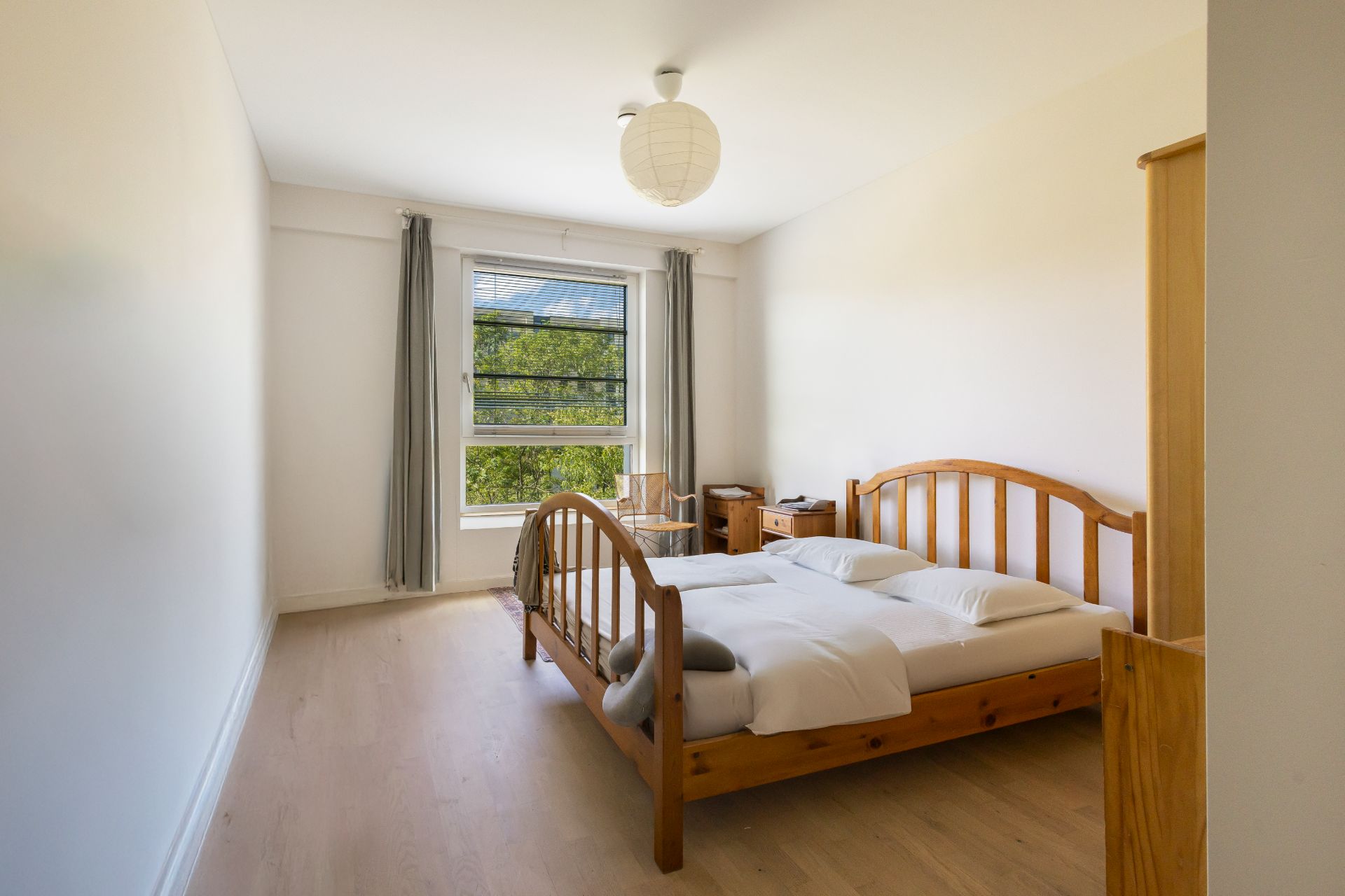 Helles Schlafzimmer mit Holzbett, Kleiderschrank, Schreibtisch und großem Fenster mit Vorhängen.