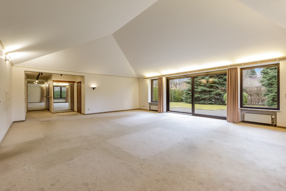 Großes, leeres Wohnzimmer mit Teppichboden und großen Fenstern mit Blick auf den Garten.