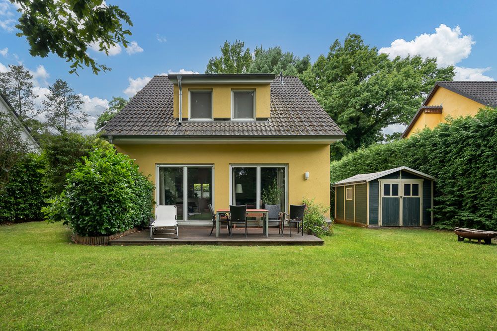 Gelbes Einfamilienhaus mit Terrasse, Gartenmöbeln und Gartenhaus im grünen Garten.