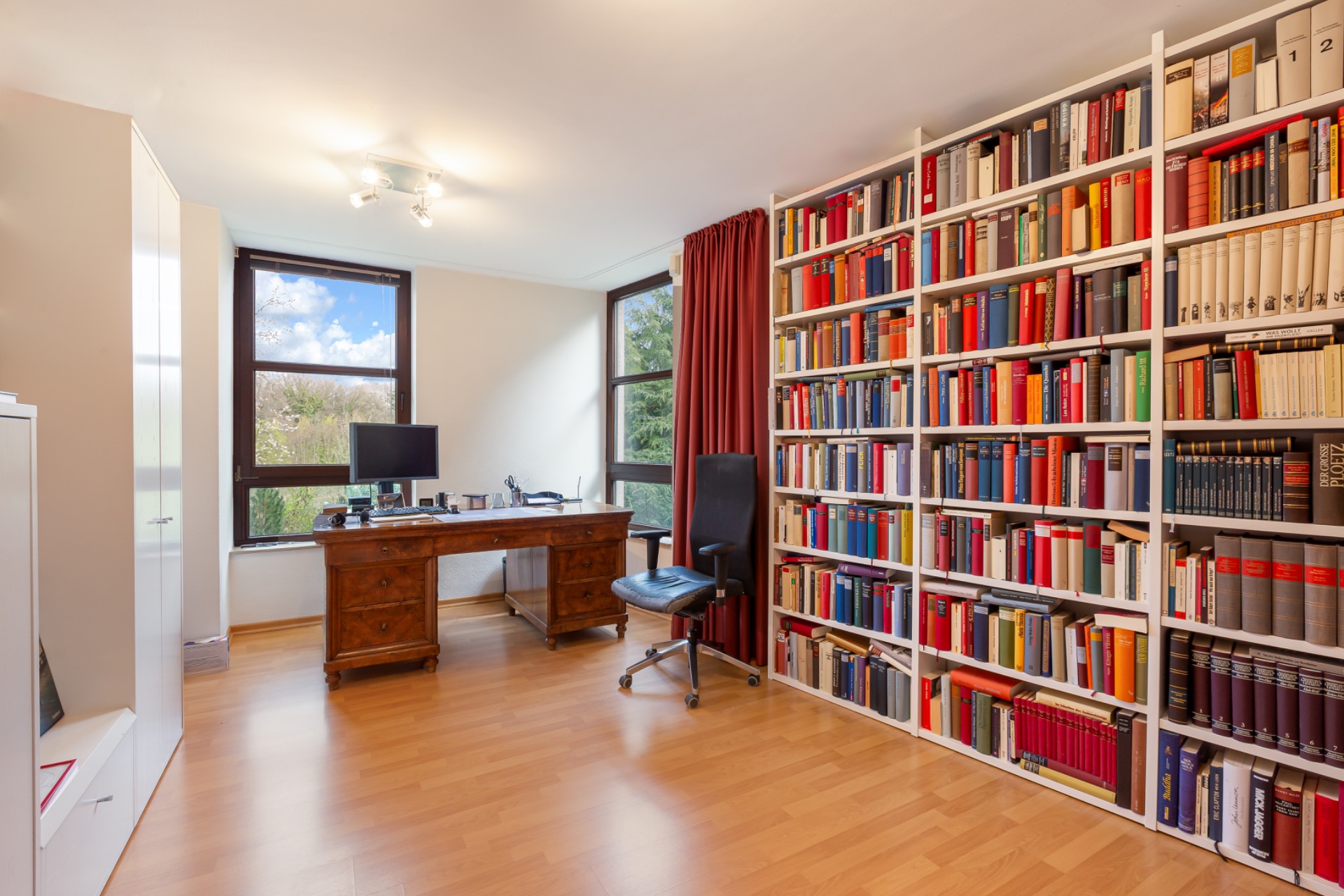 Helles Arbeitszimmer mit großem Bücherregal, Schreibtisch, Stuhl und zwei Fenstern.