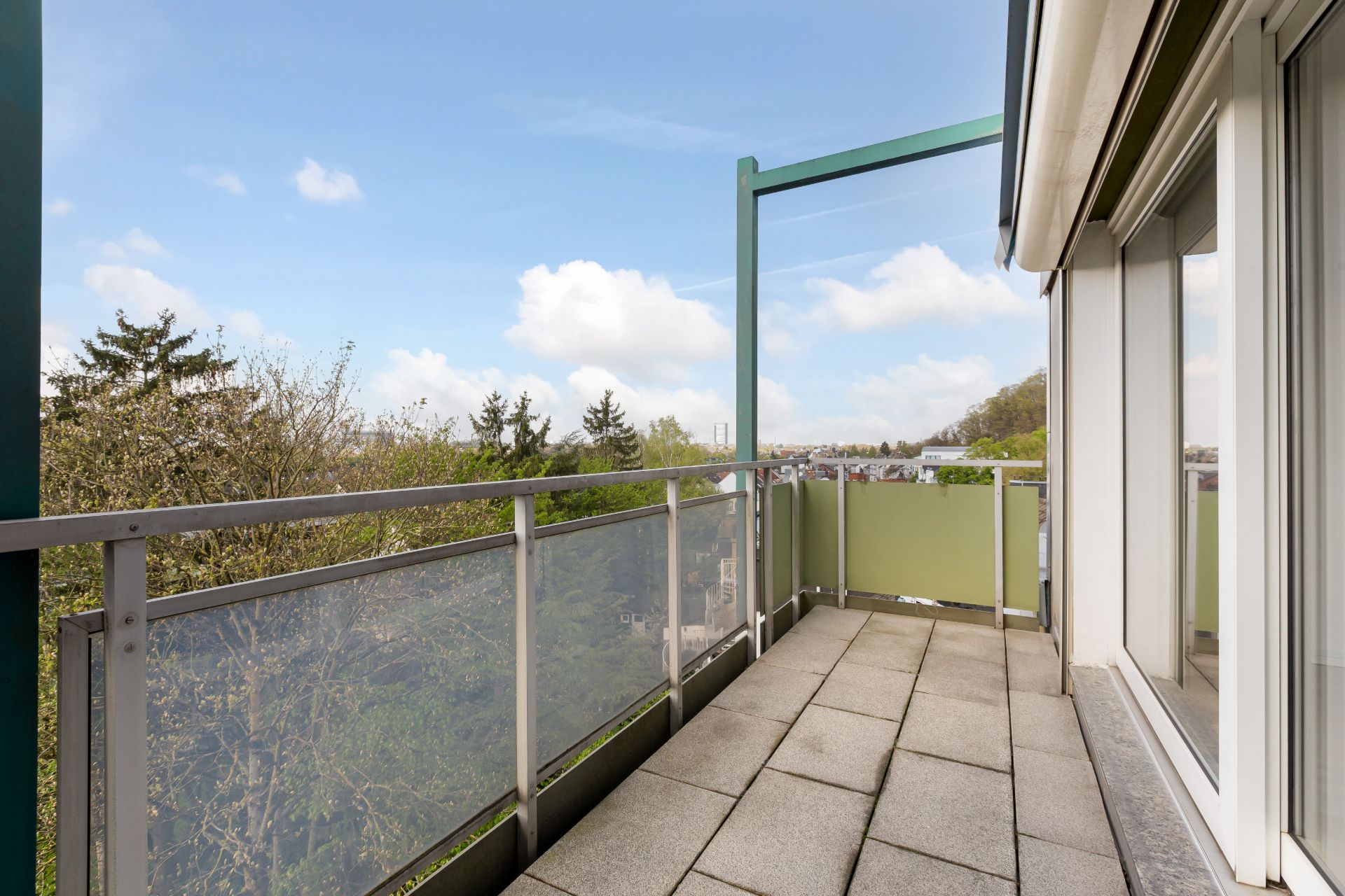 Balkon mit Glasgeländer und Fliesenboden, Blick auf Bäume und Himmel.