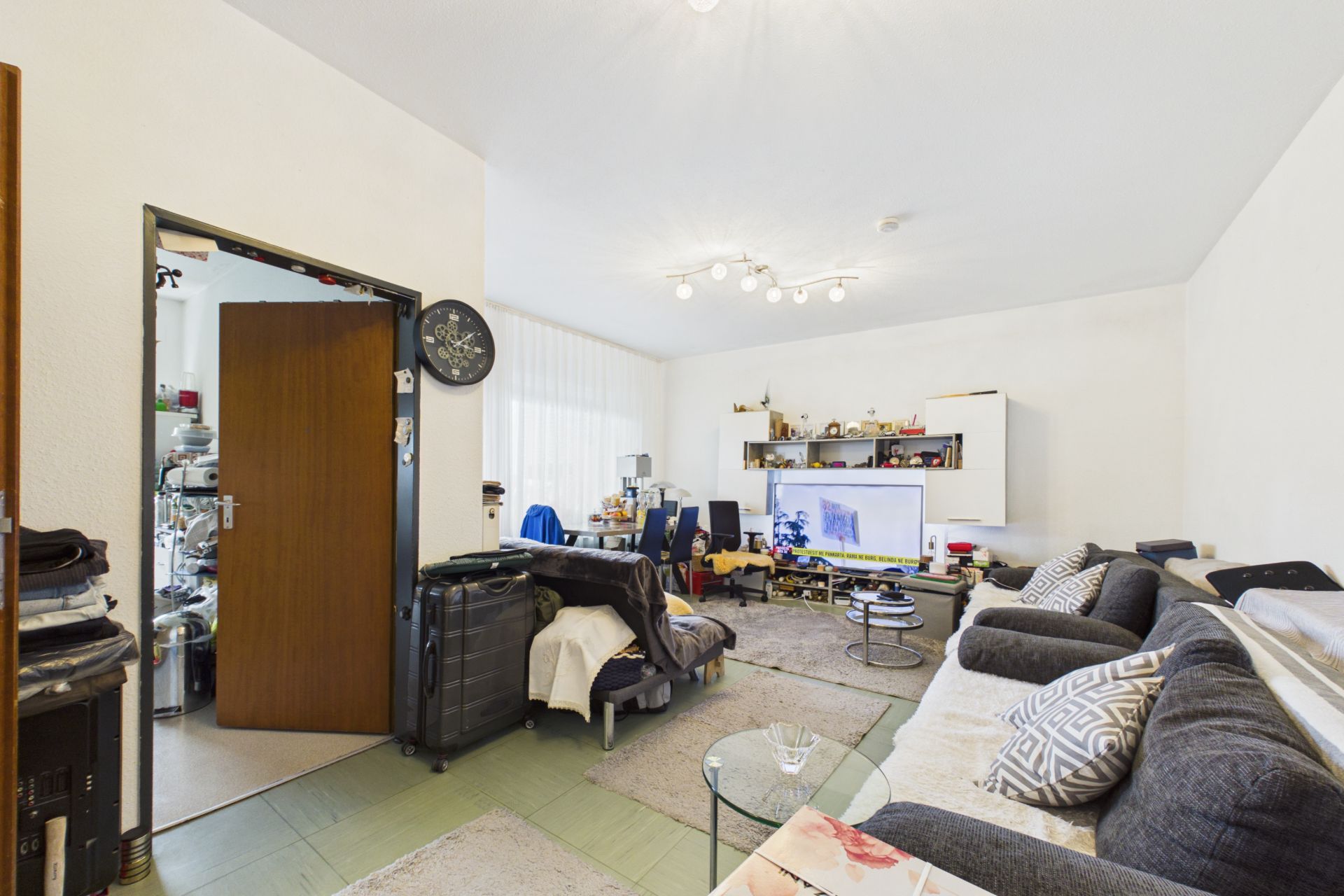 Wohnzimmer mit zwei Sofas, Glastisch, Fernseher, Regalen und offenem Durchgang zur Küche.