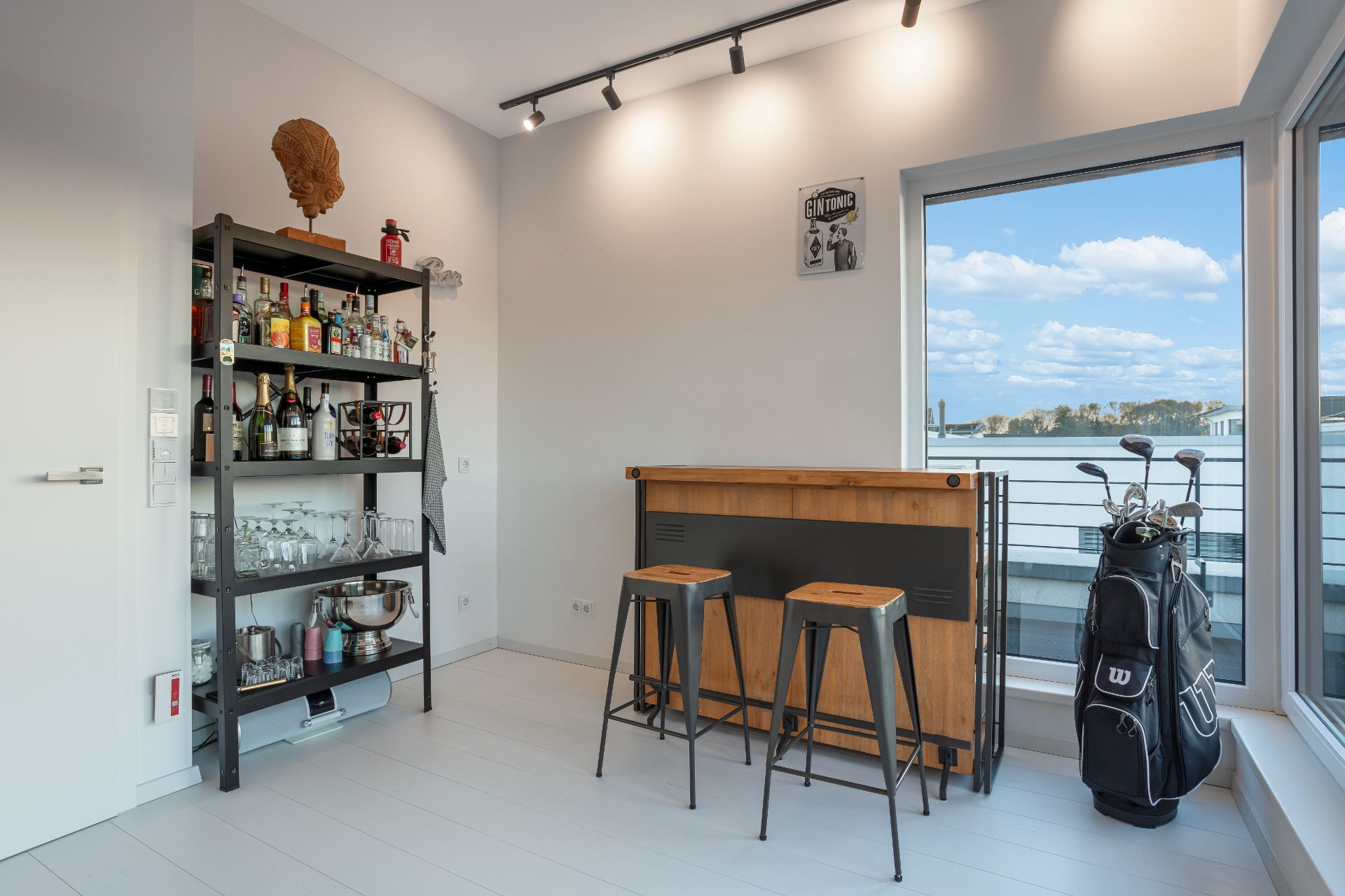 Heller Raum mit Bar, zwei Hockern, Regal mit Flaschen und Gläsern sowie Golfschlägern in einer Tasche.