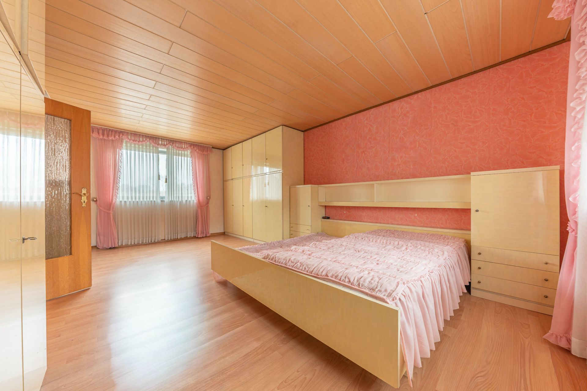 Schlafzimmer mit großem Bett, Einbauschränken und rosa Vorhängen an den Fenstern.