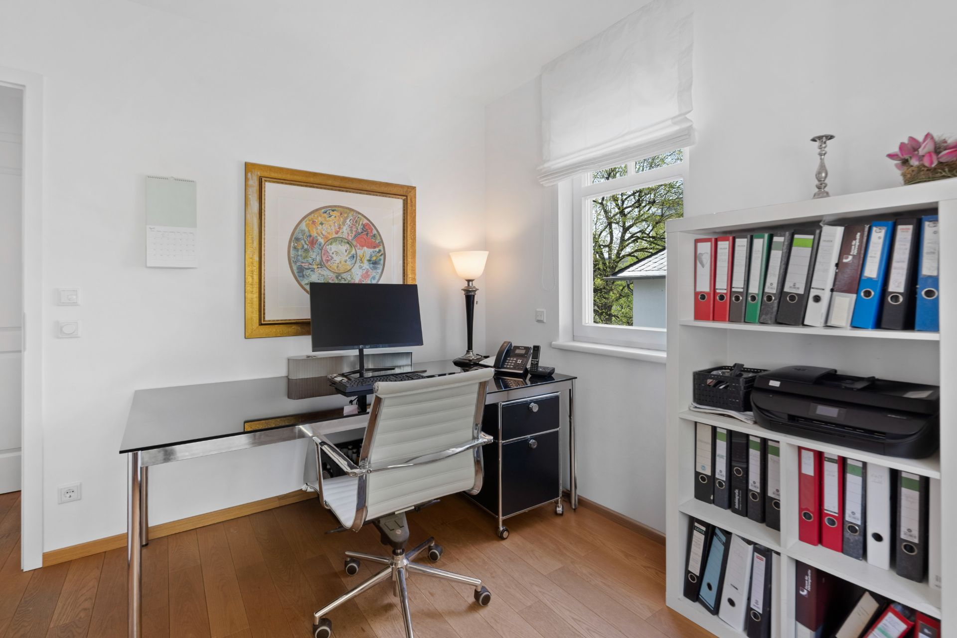 Helles Arbeitszimmer mit Schreibtisch, Computer, Regal mit Ordnern und Fenster.
