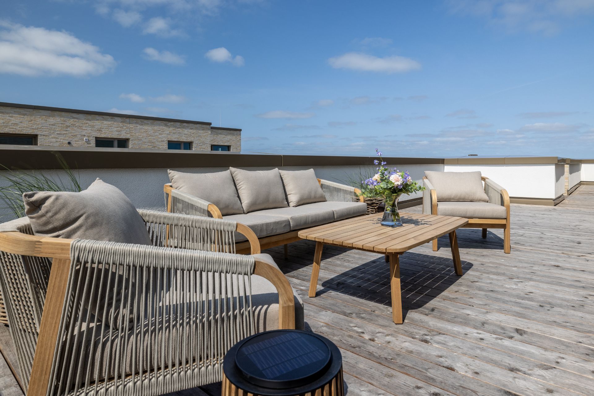 Möblierte Dachterrasse mit Holzboden, Sitzgruppe und Tisch, darauf eine Blumenvase.