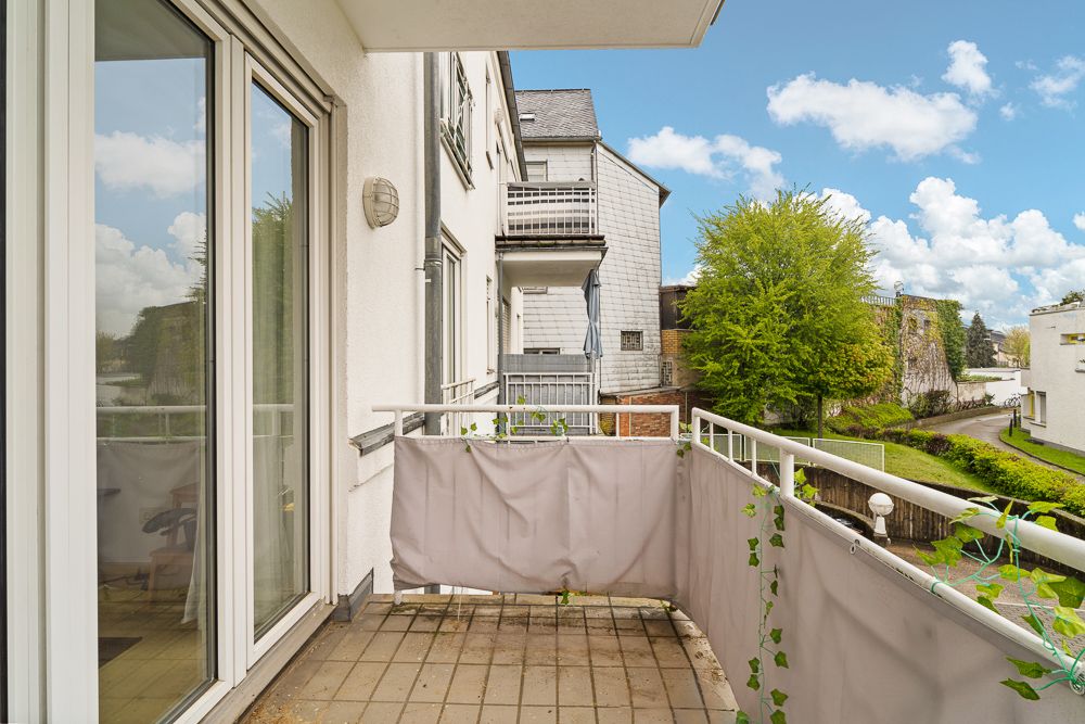 Balkon mit Glasgeländer, Blick auf Nachbarhäuser und grüne Bäume an einem sonnigen Tag.