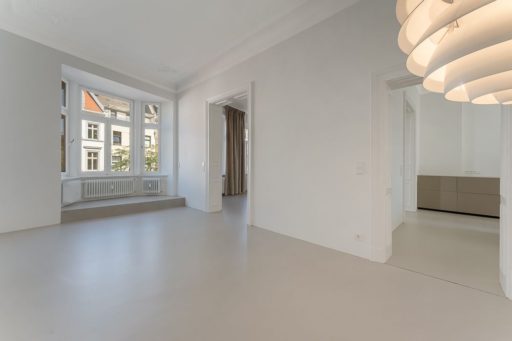 Helles, leeres Wohnzimmer mit großen Fenstern, Heizkörpern und modernem Hängelicht.